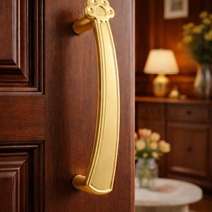 May include: A gold-colored door handle with a paw-shaped design at the top. The handle is curved and mounted on a dark brown wooden door. The background shows a glimpse of a room with a lamp and flowers.