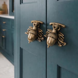 Op de afbeelding: Close-up van twee messing eikelvormige kastgrepen gemonteerd op een teal-gekleurde kastdeur. De eikelgrepen hebben ingewikkelde details en zijn symmetrisch geplaatst. De kastdeur heeft een verzonken paneelontwerp, wat diepte toevoegt.