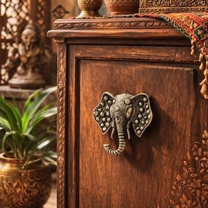 Puede incluir: Un mueble de madera oscura con un tirador de puerta de cabeza de elefante de latón adornado. El mueble tiene detalles florales tallados y está rematado con objetos decorativos, incluyendo un pequeño cofre y un textil estampado.