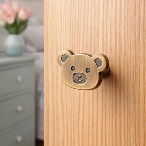 Könnte beinhalten: Ein bronzefarbener Schrankknauf in Form eines Teddybärgesichts. Das Gesicht des Bären hat dunkelgraue Akzente für Ohren, Augen und Nase. Der Knauf ist an einer hellen Holztür befestigt.
