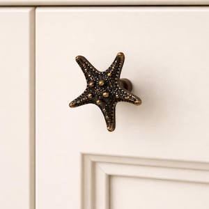 May include: Bronze starfish cabinet knob. The five-pointed knob is mounted on a white cabinet door. The knob has a textured surface with small, raised dots. The hardware adds a coastal or nautical theme to furniture.