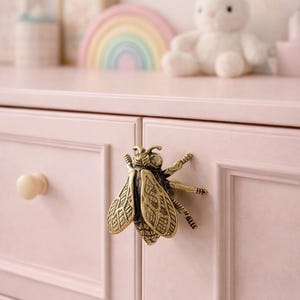Op de afbeelding: Een roze dressoir met een decoratieve messing bij-vormige handgreep. De dressoir heeft ook een crèmekleurige knop en een blauwe knop. Een knuffelkonijn en een regenboogdecoratie zijn zichtbaar op de achtergrond.