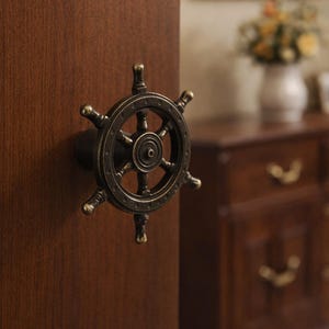 Pode incluir: Um puxador de armário bronzeado em forma de leme de navio está montado em uma porta de armário de madeira marrom escuro. O puxador tem um design circular com raios e um cubo central. O fundo mostra uma cômoda de madeira desfocada e uma imagem emoldurada.