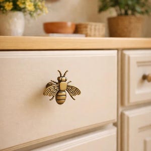 Puede incluir: Primer plano de un cajón de armario color crema con un tirador decorativo en forma de abeja de latón. El tirador está detallado con alas y segmentos corporales. El armario forma parte de un conjunto de cocina o baño.