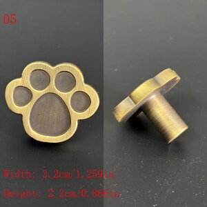 Puede incluir: Tirador de armario con huella de pata de lat&oacute;n antiguo. El tirador tiene forma de pata de perro o gato con una zona central rebajada. La anchura es de 3,2 cm y la altura de 2,2 cm. El n&uacute;mero "05" est&aacute; en la esquina superior izquierda.
