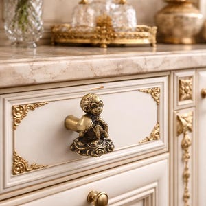 May include: Close-up of a white cabinet with gold accents and a decorative monkey-shaped drawer pull. The cabinet has ornate gold trim and a marble countertop. Other decorative items are in the background.