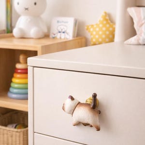 May include: A cream-colored dresser drawer with a cat-shaped knob. The knob is white and brown with a gold-colored base. A white bunny lamp, a star-shaped pillow, and a stack of books are visible in the background.