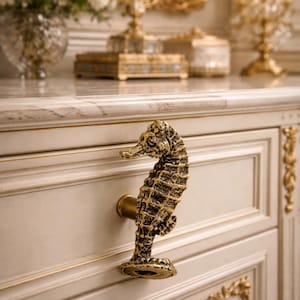 Puede incluir: Tirador decorativo con forma de caballito de mar, acabado en oro antiguo. El caballito de mar está montado en una puerta de armario crema con detalles dorados. El diseño añade un toque de elegancia al mueble.