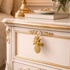 Può includere: Comodino color crema con finiture dorate e pomello a forma di ananas. Il cassetto è incorniciato da dettagli dorati e il comodino presenta dettagli dorati ornati. Una lampada dorata e un vaso di fiori bianchi sono visibili.
