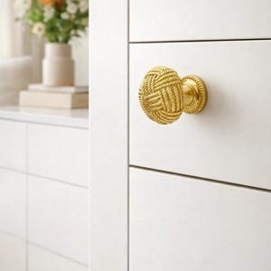 May include: A gold-colored cabinet knob with a woven rope design. The knob is round and attached to a white cabinet door. The knob's design is a prominent feature, adding a decorative element to the cabinet. The image also shows a vase of flowers.