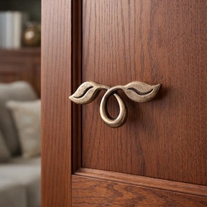 Puede incluir: Manija de puerta en tono bronce con diseño de hojas. La manija está montada en una puerta de madera marrón oscuro, con vetas visibles. El diseño de la manija presenta dos hojas estilizadas conectadas a un tirador circular.
