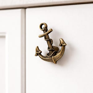 Peut inclure: Poignée de meuble en forme d'ancre de couleur bronze. La quincaillerie sur le thème nautique présente un design détaillé avec une corde enroulée autour de la barre transversale de l'ancre. L'ancre est montée sur une porte d'armoire blanche.