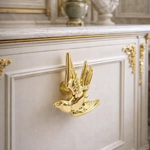 Puede incluir: Un tirador de armario dorado con forma de pájaro está montado en una puerta de armario blanca. El pájaro está en vuelo, con las alas extendidas. El armario tiene adornos dorados y forma parte de un mueble más grande.