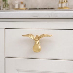 Op de afbeelding: Goudkleurige adelaarvormige kastgreep op een witte lade. De greep heeft uitgespreide vleugels en een bolvormige basis, wat een decoratieve touch geeft aan keuken- of badkamerkasten. De hardware is helder, metallic goud.