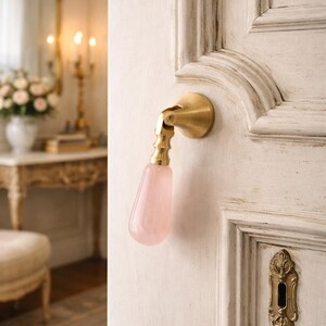 May include: A door handle with a brass base and a pink, teardrop-shaped crystal handle. The handle is mounted on a white door with decorative molding and a brass keyhole cover. The overall aesthetic is vintage and elegant.