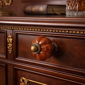 Può includere: Primo piano di un cassetto di cassettiera in legno marrone scuro con una manopola decorativa a forma di zucca. La manopola è di un marrone caldo e lucido con una base color ottone. Il cassetto fa parte di un mobile più grande con accenti dorati.