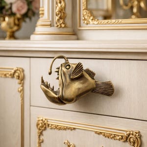 Könnte beinhalten: Messingfarbener Schrankgriff in Form eines Anglerfischs. Der Griff ist an einem hellen Schrank mit goldenen Zierleisten angebracht. Der Schrank ist Teil eines größeren Möbelstücks mit aufwendigen Details und einem Blumenarrangement im Hintergrund.