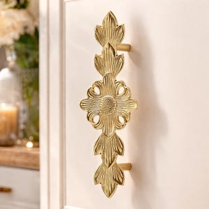 Op de afbeelding: Goudkleurige kastgreep met een sierlijk bloemmotief. De greep heeft een centrale bloemvorm met gedetailleerde bloemblad- en bladeraccenten. De hardware is gemonteerd op een witte kastdeur.