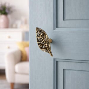 May include: A brass leaf-shaped door handle is mounted on a light blue door. The handle has intricate detailing, resembling the veins of a leaf. The door is part of a room with a soft, neutral color palette.