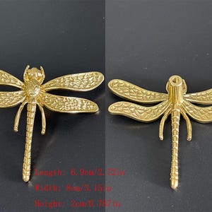 Puede incluir: Adorno de lib&eacute;lula dorado. La lib&eacute;lula tiene alas detalladas y un cuerpo segmentado. La longitud es de 6,9 cm, la anchura es de 8 cm y la altura es de 2 cm. La lib&eacute;lula se muestra sobre un fondo oscuro.