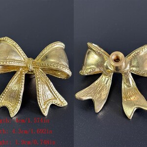 Puede incluir: Dos piezas de herrajes decorativos dorados en forma de lazo. Los herrajes miden 4 cm de largo, 4,3 cm de ancho y 1,9 cm de alto. Los herrajes tienen un orificio roscado en la parte posterior. El texto de la imagen dice: Longitud: 4 cm, Ancho: 4,3 cm, Altura: 1,9 cm.