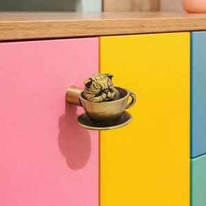 Può includere: Pomello per armadio in bronzo a forma di gatto addormentato in una tazza da tè. Il pomello è montato su un'anta di un armadio rosa e giallo. L'impugnatura della tazza è sul lato sinistro.