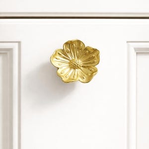 Puede incluir: Un pomo de armario dorado con forma de flor. El pomo está montado en una puerta de armario blanca. La flor tiene pétalos detallados y un centro texturizado, lo que añade un toque de elegancia al armario.