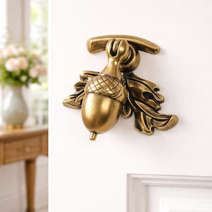 Op de afbeelding: Een messing deurklopper in de vorm van een eikel, met gedetailleerde bladeren. De klopper is gemonteerd op een witte deur, wat het ingewikkelde ontwerp en de warme metalen afwerking benadrukt. De dop van de eikel heeft een textuur.