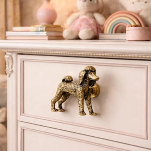 May include: A light pink dresser with a decorative brass poodle drawer pull. The dresser has two drawers with a smaller, round knob on the bottom drawer. A lamp, stuffed bunny, and rainbow decor are on top.