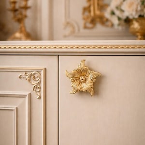 Pode incluir: Close-up de uma porta de armário creme com acabamentos dourados e uma puxador decorativo dourado em forma de flor. O armário tem detalhes ornamentados e faz parte de uma peça de mobiliário maior.