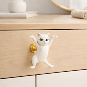 以下が含まれることがあります： 白い猫の形をしたキャビネットノブ。腕を広げ、緑色の目をしています。ノブは明るい木製の引き出しに取り付けられており、金色のベースが付いています。猫は家具の気まぐれな装飾要素です。