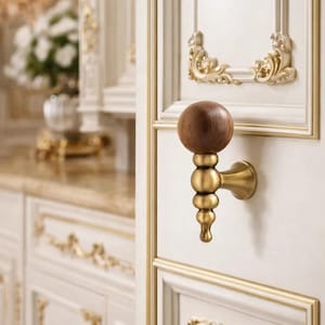 May include: Close-up of a wooden and brass cabinet knob. The knob has a dark brown, spherical handle atop a brass stem with a series of rounded sections. The cabinet door is white with gold trim and decorative accents. The overall theme is luxury.