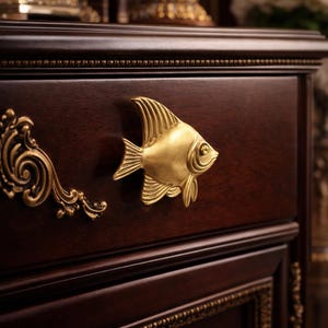 May include: Close-up of a dark brown wooden cabinet drawer with a gold-colored fish-shaped handle. The drawer has ornate gold trim and a decorative gold accent on the left side. The cabinet is part of a larger furniture piece.