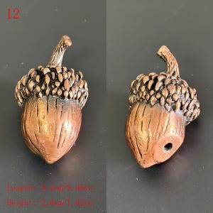 Puede incluir: Dos esculturas de bellotas de color bronce. Las tapas de las bellotas son texturizadas y de color marr&oacute;n oscuro, mientras que los cuerpos de las bellotas son lisos y m&aacute;s claros. El texto "12" est&aacute; en la esquina superior izquierda. Longitud 5,2 cm, altura 2,6 cm.