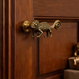 Może przedstawiać: Brązowa klamka do drzwi w kształcie kameleona zamontowana na ciemnobrązowych drewnianych drzwiach. Klamka jest misternie zdobiona, przedstawiając cechy gada. Drzwi mają klasyczny wzór paneli.