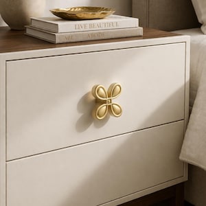 Op de afbeelding: Een crèmekleurig nachtkastje met een donkerhouten blad en voet. Het heeft een goudkleurige bloemvormige ladeknop met vier bloemblaadjes. Bovenop liggen boeken en een goudkleurige kom. De boeken tonen de woorden "LIVE BEAUTIFUL" en "ABODE".