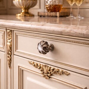 Può includere: Mobile color crema con finiture dorate e pomello decorativo argentato. Un accento floreale dorato è sotto il pomello. Un piano di lavoro in marmo ospita un vaso dorato di fiori bianchi, una caraffa e calici.
