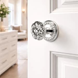May include: A silver-toned door knob with a floral design and a ring of clear crystals. The knob is mounted on a white door, with a glimpse of a bright, well-lit room in the background.