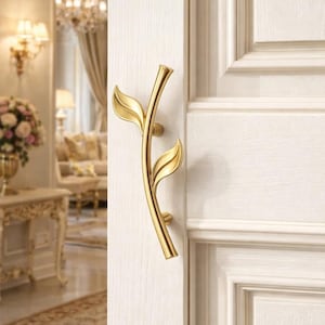 May include: A gold-colored door handle in a leaf design is mounted on a white door. The handle has a curved stem with two leaf-shaped accents. The door is part of a room with a classic, elegant aesthetic.