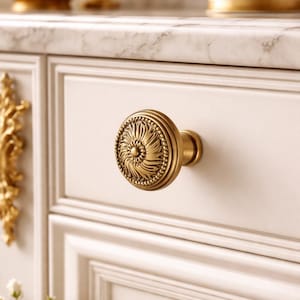 May include: Close-up of a decorative brass drawer pull with a floral design. The knob is round with a detailed, textured surface and a warm, antique gold finish. The drawer is white with a marble countertop.