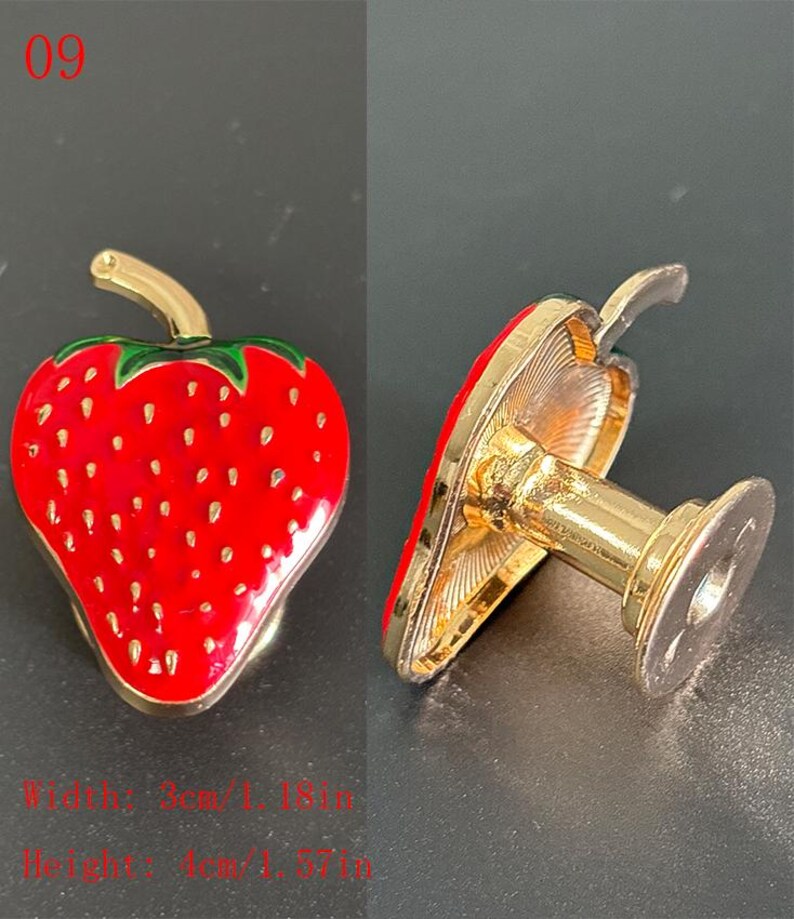 Puede incluir: Un broche decorativo con forma de fresa con acabado esmaltado rojo y detalles dorados. El broche mide 3 cm de ancho y 4 cm de alto. El dise&ntilde;o incluye hojas verdes y peque&ntilde;os puntos blancos.