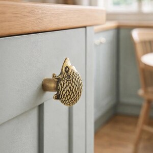 Op de afbeelding: Een messing kastknop in de vorm van een egel bevestigd aan een lichtgrijze kastdeur. De knop heeft een getextureerd lichaam en een gedetailleerd gezicht. De keukenkast heeft een houten werkblad.