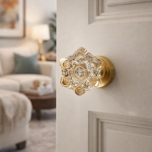 May include: A clear, faceted glass door knob with a floral design and a brass base. The knob is mounted on a white door, showcasing its intricate details and reflective qualities. The knob's dimensions are approximately 3 inches in diameter.