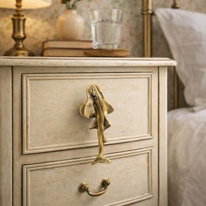 May include: A cream-colored nightstand with a decorative brass fish-shaped drawer pull. The nightstand has two drawers and a curved bottom edge. A brass lamp and a vase of flowers are on top.