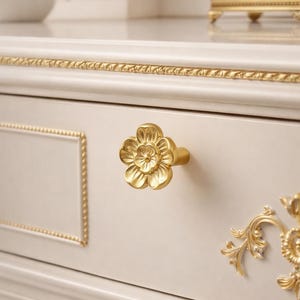Puede incluir: Primer plano de un cajón color crema con un pomo dorado en forma de flor. El cajón tiene ribetes y detalles dorados. Un elemento decorativo dorado está en el lado derecho del cajón.