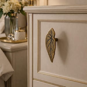 May include: Close-up of a cream-colored cabinet door with a leaf-shaped brass handle. The cabinet has gold trim and a textured surface. A lamp and vase of flowers are visible in the background.