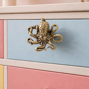 May include: A brass octopus drawer pull is fixed to a light blue drawer. The octopus has detailed tentacles and a textured head. Other drawers are pink and yellow with matching brass knobs. The furniture has a cream-coloured frame.