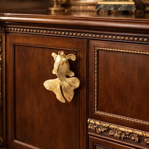 May include: A dark brown wooden cabinet with gold accents and a decorative gold fish-shaped handle. The cabinet has ornate detailing and a classic design, with a candle and clock in the background.