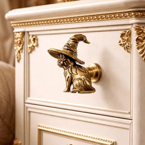 May include: A bronze-colored cabinet knob shaped like a dog wearing a witch hat. The knob is mounted on a cream-colored cabinet with gold trim and decorative accents. The cabinet has two drawers.