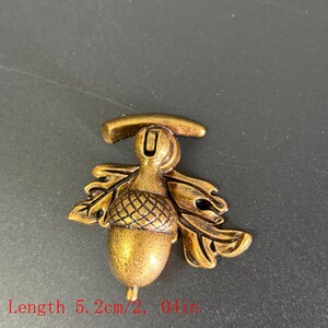 Puede incluir: Elemento decorativo en forma de bellota de color bronce. La bellota mide 5,2 cm de largo, 5,1 cm de ancho y 2,2 cm de alto. El dise&ntilde;o incluye hojas de roble y el n&uacute;mero "02" en la esquina superior izquierda.
